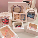 An array of beautifully decorated decoden items by Deco Deco, each displayed in individual white boxes with viewing windows. The collection includes custom-decorated items such as mirrors, frames, and cases, each intricately adorned with charms, glitter, and vibrant designs, presented in a soft pink setting with the Deco Deco logo visible.