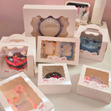 An array of beautifully decorated decoden items by Deco Deco, each displayed in individual white boxes with viewing windows. The collection includes custom-decorated items such as mirrors, frames, and cases, each intricately adorned with charms, glitter, and vibrant designs, presented in a soft pink setting with the Deco Deco logo visible.