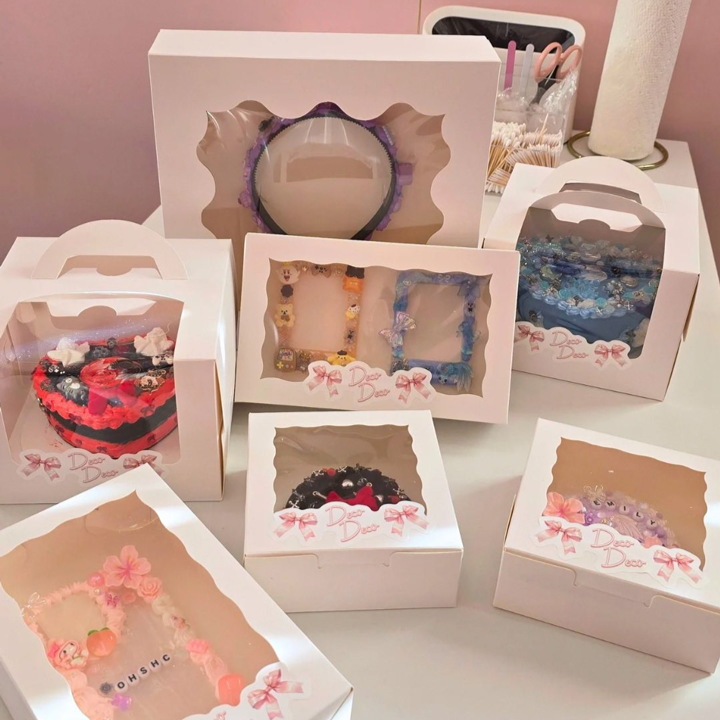 An array of beautifully decorated decoden items by Deco Deco, each displayed in individual white boxes with viewing windows. The collection includes custom-decorated items such as mirrors, frames, and cases, each intricately adorned with charms, glitter, and vibrant designs, presented in a soft pink setting with the Deco Deco logo visible.
